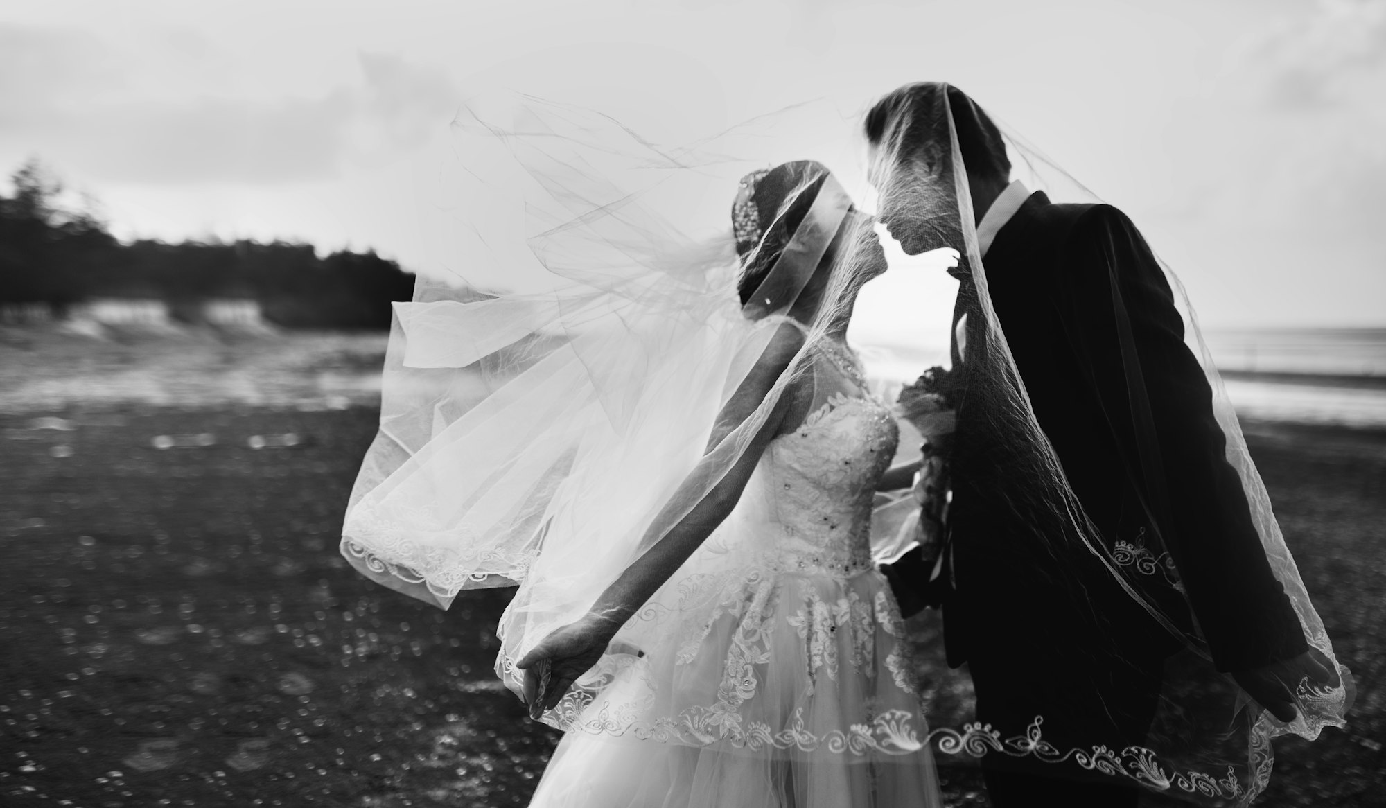 Black and white photo of a couple kissing under a wind-blown veil